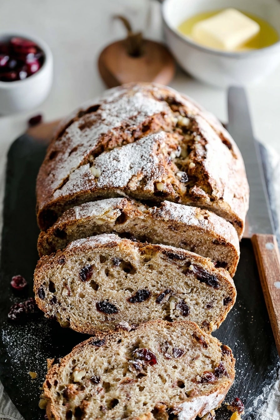 A rustic loaf of cranberry walnut bread, dusted with flour and sliced on a dark slate board.