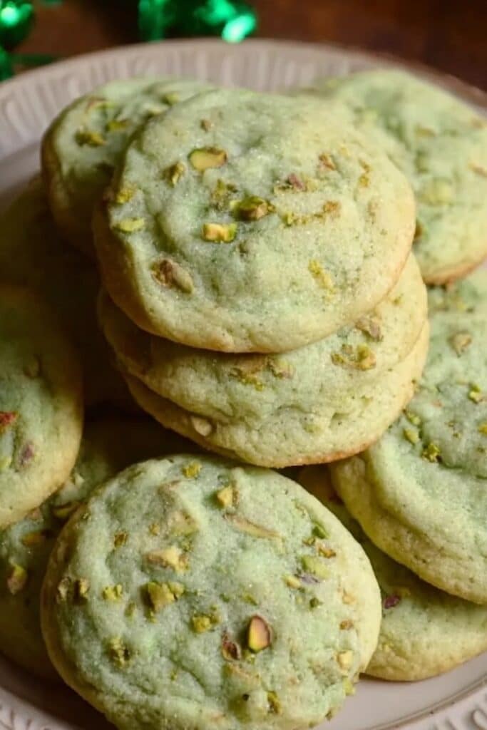 A close-up of a plate piled high with soft, pale green pistachio pudding cookies, with visible chunks of chopped pistachios on top.
