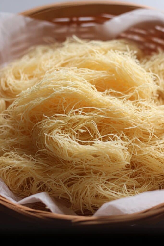 A close-up of fresh, uncooked homemade kataifi dough, showing its distinctive thin strands resting in a parchment-lined basket.