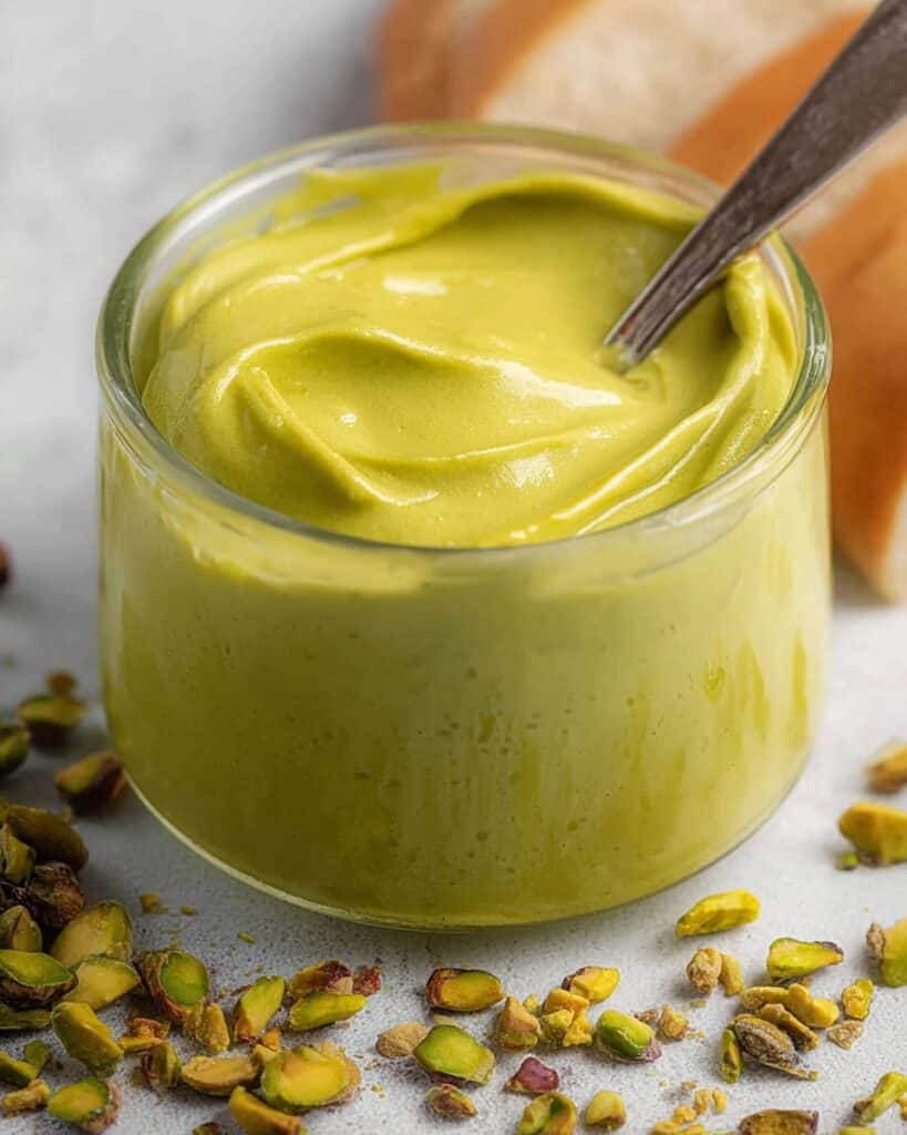 A close-up shot of a small glass jar filled with creamy, vibrant green pistachio cream. A small spoon is dipped into the cream, and shelled pistachios are scattered around the base of the jar on a light-colored surface.