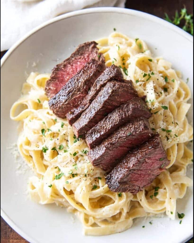 A close-up overhead shot of a white bowl filled with creamy fettuccine Alfredo, topped with several slices of perfectly cooked medium-rare steak fanned out, garnished with fresh parsley and grated Parmesan cheese
