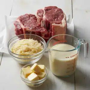 Ingredients for steak fettuccine alfredo laid out on a white wooden surface: two raw New York strip steaks on parchment paper, a glass bowl of grated Parmesan cheese, a measuring jug of heavy cream, and a small glass bowl of butter cubes