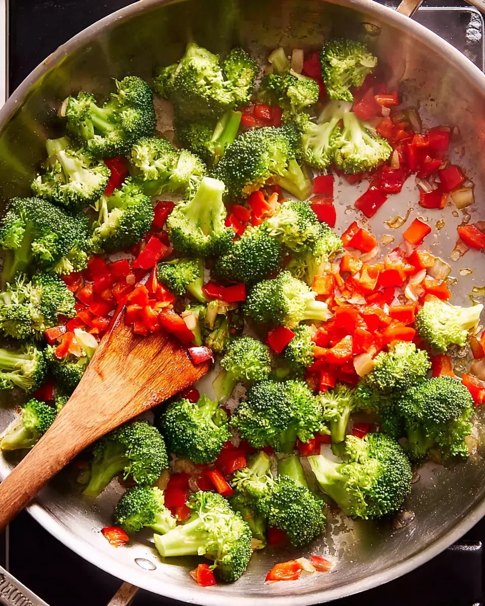 Cook the Vegetables An overhead shot of bright green broccoli florets and diced red bell peppers being stir-fried in a large skillet with a wooden spoon.