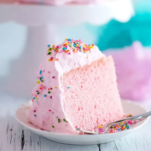 A vertical close-up of a single slice of fluffy pink angel food cake on a white scalloped plate. The slice features a light pink airy crumb, billowy pink marshmallow frosting, and is topped with rainbow sprinkles. A silver fork rests on the plate beside the cake.