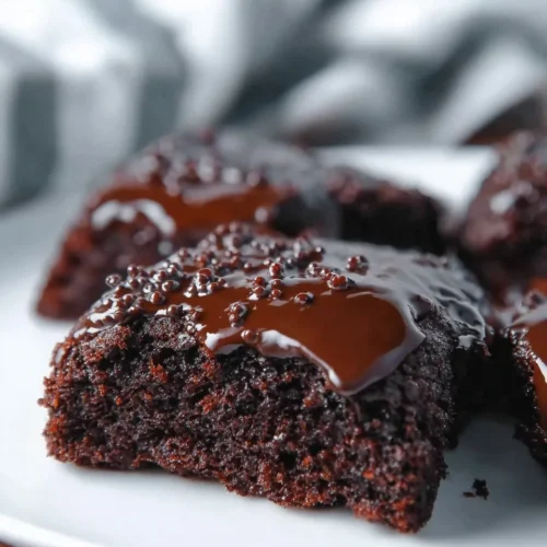 A macro close-up shot of a moist Midnight Fudge Cake square resting on a white plate. The cake is topped with a layer of glossy dark chocolate ganache and scattered with small chocolate sprinkles.
