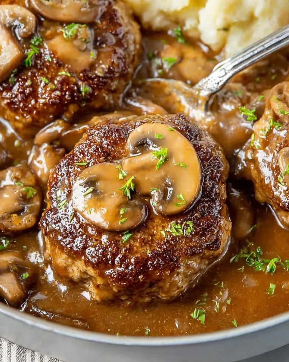 juicy Salisbury steak patty topped with a large mushroom Close-up of a juicy Salisbury steak patty topped with a large mushroom slice and rich brown gravy, garnished with fresh parsley, with a spoon serving the sauce next to mashed potatoes.