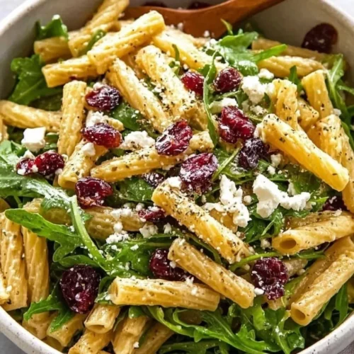 Overhead close-up view of Feta and Cranberry Rigatoni Salad in a speckled ceramic bowl, featuring rigatoni pasta, fresh spinach leaves, dried cranberries, and crumbled feta cheese with a wooden spoon tucked into the side.