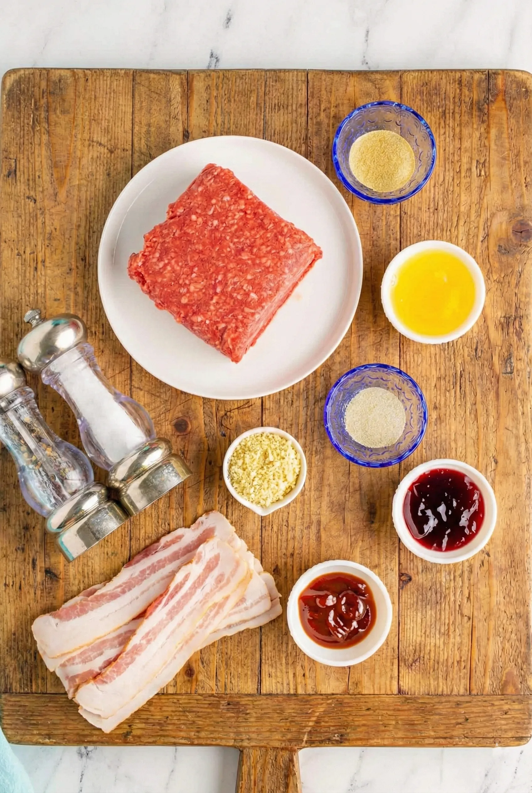 Ingredients-Bacon-Wrapped-Meatball Overhead flat lay of ingredients for Cheddar Stuffed BBQ Bacon Bombs on a wooden board, featuring raw ground beef on a plate, slices of bacon, breadcrumbs, beaten egg, seasonings in small bowls, salt and pepper grinders, and BBQ sauce.