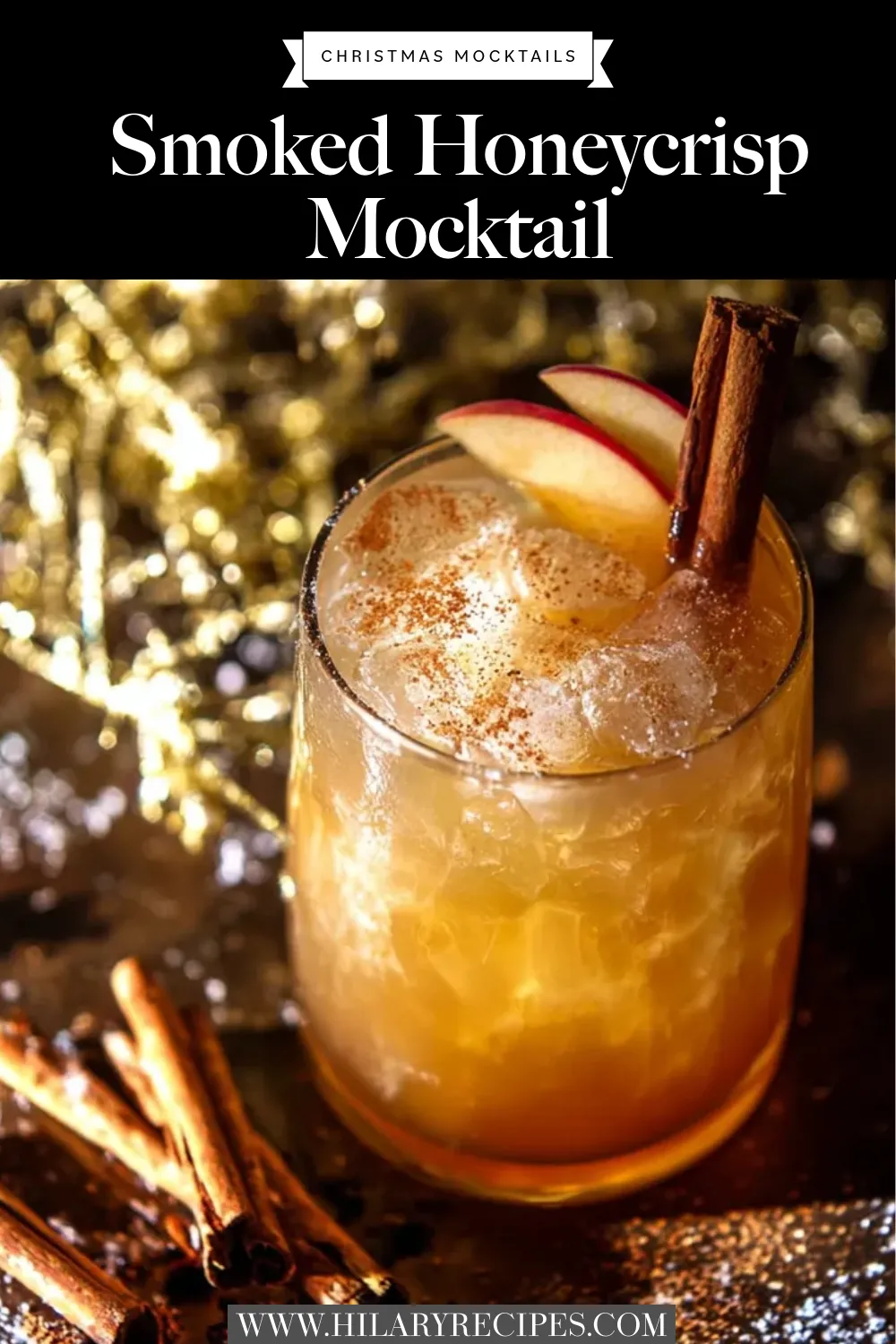 A Smoked Honeycrisp Mocktail in a glass with ice, garnished with fresh apple slices and cinnamon sticks, against a festive gold bokeh background.