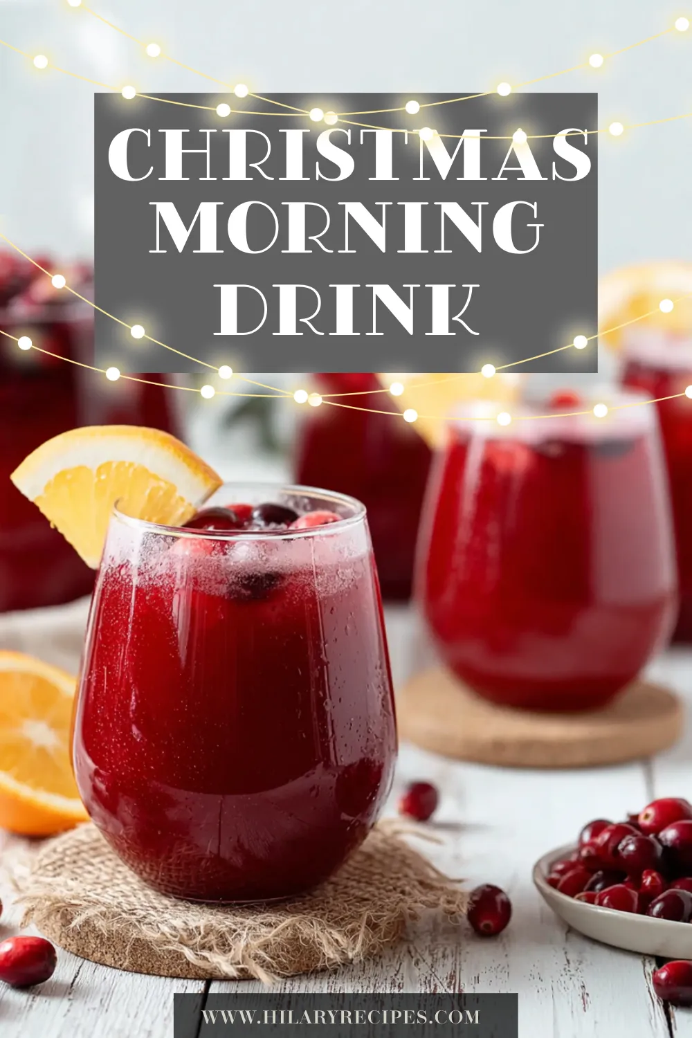 A stemless glass filled with a vibrant red Christmas morning drink, garnished with an orange slice and fresh cranberries. The glass sits on a burlap coaster, with more glasses of the non-alcoholic punch and a pitcher visible in the festive background.