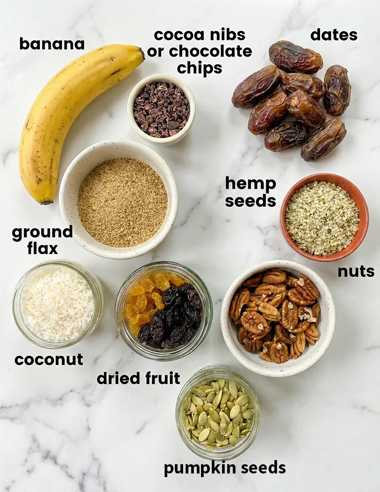 ingredients for nut and seed energy cookies An overhead flat lay photo showing the ingredients for nut and seed energy cookies labeled with text. The spread includes a ripe banana, Medjool dates, pumpkin seeds, pecans, hemp seeds, ground flax, dried fruit, shredded coconut, and cacao nibs on a white marble surface.