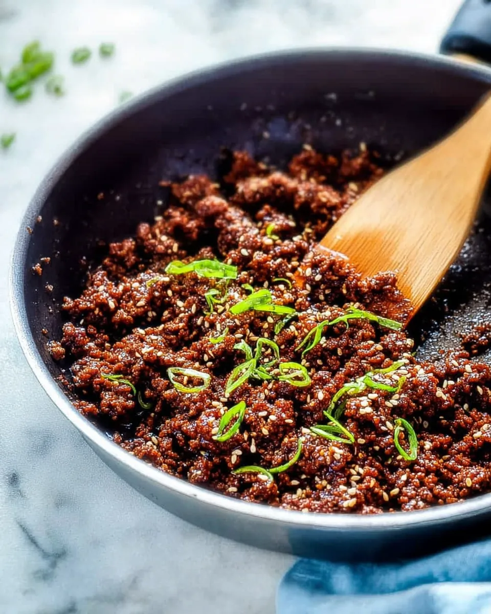 Korean BBQ ground beef cooking Overhead shot of Korean BBQ ground beef cooking in a dark skillet. The meat is coated in a thick, glossy dark brown soy glaze and generously garnished with fresh green onions and white sesame seeds. A wooden spoon rests in the pan.