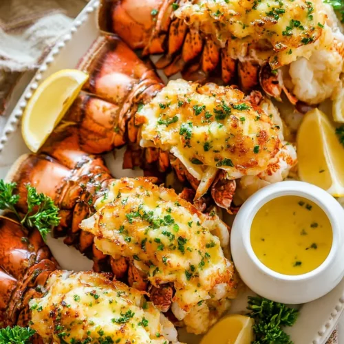 Close-up overhead shot of golden-brown baked lobster tails arranged on a white serving platter. The tails are garnished with fresh parsley and lemon wedges, sitting next to a small white bowl filled with melted garlic butter for dipping.