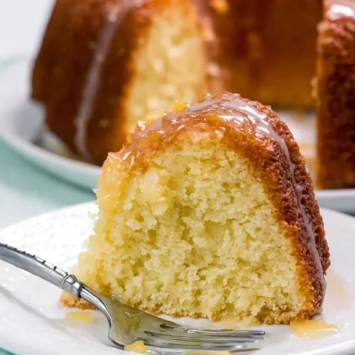 A slice of moist pineapple pound cake on a white dessert plate, topped with a glossy glaze and served with a silver fork, with the rest of the bundt cake visible in the background.