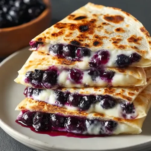 A close-up, appetizing stack of three golden-brown blueberry breakfast quesadilla wedges on a beige plate, overflowing with warm, melting cream cheese and juicy cooked blueberries, with a bowl of fresh blueberries in the background.