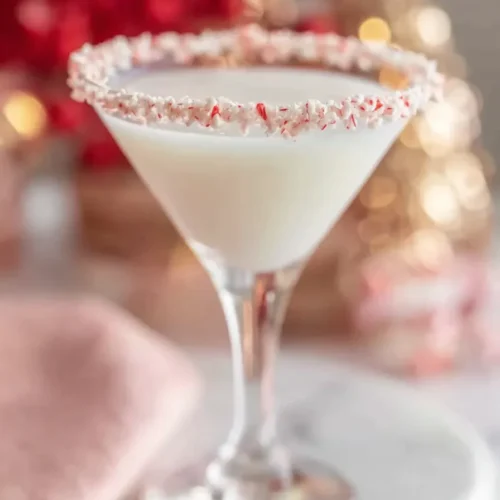 A side-angle shot of a creamy white mocktail in a martini glass, showing a thick, perfectly applied rim of crushed red and white candy canes, set against a blurred background of festive red and gold holiday lights.