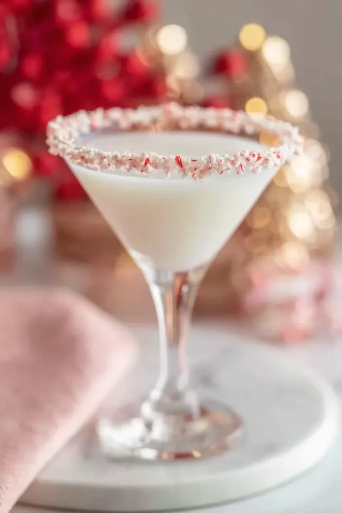 A side-angle shot of a creamy white mocktail in a martini glass, showing a thick, perfectly applied rim of crushed red and white candy canes, set against a blurred background of festive red and gold holiday lights.