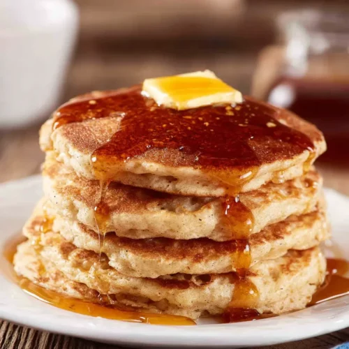 A close-up view of a stack of four fluffy Vanilla Cinnamon Buttermilk Pancakes topped with a pat of melting butter and drenched in rich maple syrup, sitting on a white plate against a rustic wooden background.