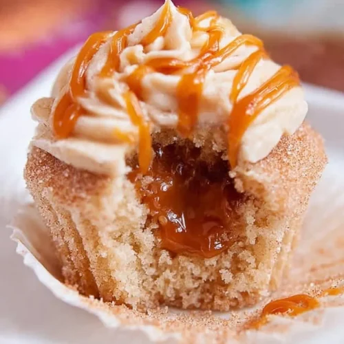 Cross-section view of a Churro Caramel Crunch Cupcake with a bite taken out, revealing the oozing homemade caramel filling center inside the cinnamon sponge.