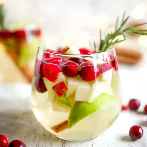 A stemless wine glass filled with sparkling non-alcoholic white sangria, garnished with fresh cranberries, diced apples, a cinnamon stick, and a sprig of rosemary.