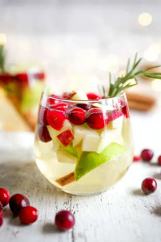 A stemless wine glass filled with sparkling non-alcoholic white sangria, garnished with fresh cranberries, diced apples, a cinnamon stick, and a sprig of rosemary.