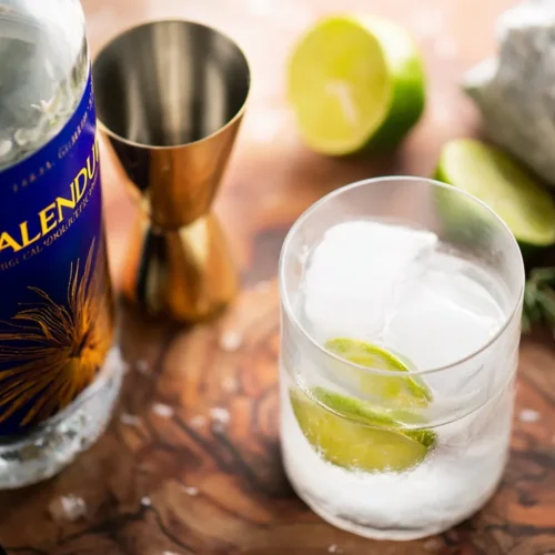 A lowball glass filled with large ice cubes, ready for a mocktail. Next to it are a bottle of Caleno non-alcoholic spirit, a metal jigger, and several lime wedges on a marble surface.