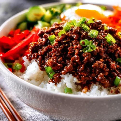 Close-up side view of a white bowl filled with fluffy rice, topped with glossy Korean BBQ ground beef, sliced red peppers, cucumbers, and a fried egg. The dish is garnished with green onions and sesame seeds, with wooden chopsticks resting on the side.