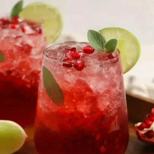 A close-up of a vibrant red pomegranate mocktail in a rounded glass, filled with crushed ice and garnished with a lime wheel, a fresh sage leaf, and scattered pomegranate arils.