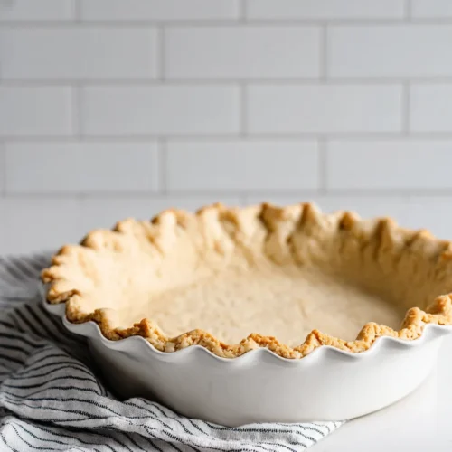 A par-baked, flaky pie crust with a ruffled edge, sitting in a white fluted pie dish. The dish rests on a black-and-white striped kitchen towel on a white counter, with a white subway tile backsplash in the background.