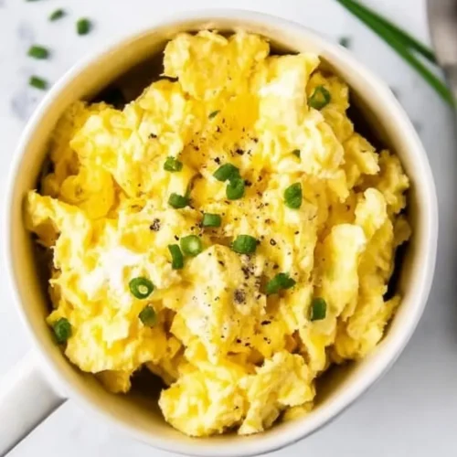Close-up overhead view of fluffy microwave scrambled eggs in a white mug, garnished with chopped fresh chives and cracked black pepper.