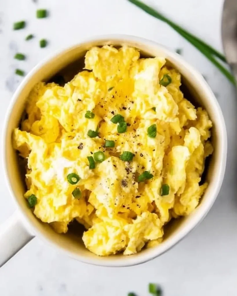 Close-up overhead view of fluffy microwave scrambled eggs in a white mug, garnished with chopped fresh chives and cracked black pepper.
