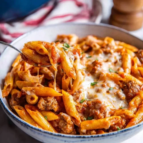 Close-up of creamy One-Pot Pasta with Sausage for Two in a bowl, featuring a spoonful of cheesy penne pasta with meat sauce being lifted to show a cheese pull, garnished with fresh herbs.