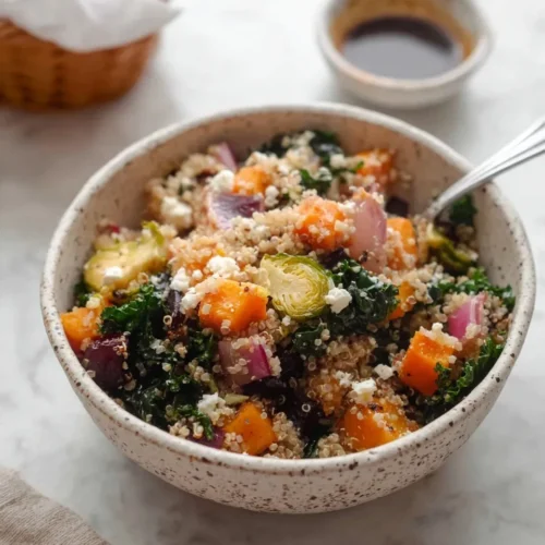 Close-up of a speckled ceramic bowl filled with Quinoa Salad with Roasted Vegetables, featuring roasted butternut squash, beets, Brussels sprouts, kale, and crumbled feta, with a small bowl of balsamic dressing in the background.