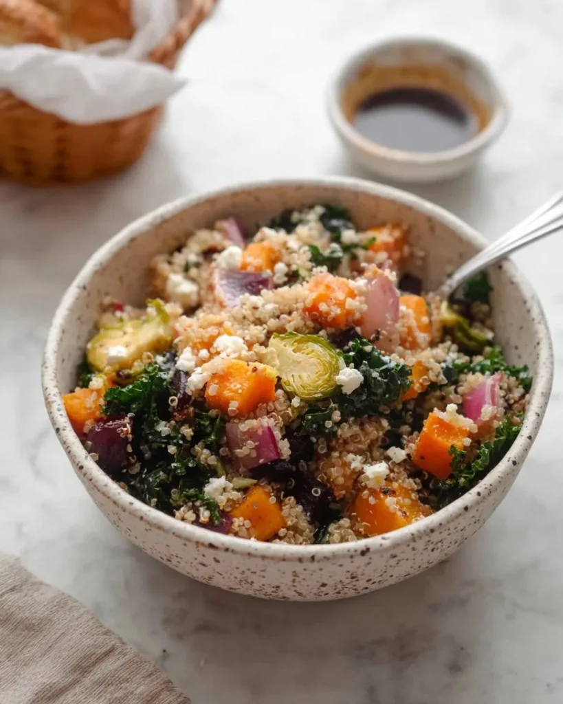 Close-up of a speckled ceramic bowl filled with Quinoa Salad with Roasted Vegetables, featuring roasted butternut squash, beets, Brussels sprouts, kale, and crumbled feta, with a small bowl of balsamic dressing in the background.
