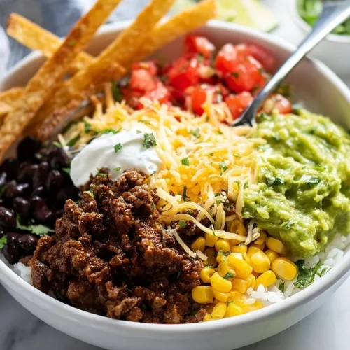 A close-up, high-angle shot of a white bowl filled with a colorful bean and rice burrito bowl, featuring sections of seasoned ground beef, black beans, corn, diced tomatoes, shredded cheese, guacamole, sour cream, and crispy tortilla strips.