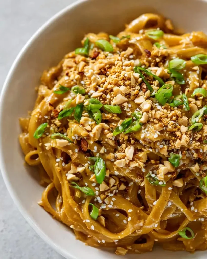 Close-up overhead shot of creamy peanut noodles in a white bowl, heavily garnished with chopped peanuts, sliced green onions, sesame seeds, and red pepper flakes.