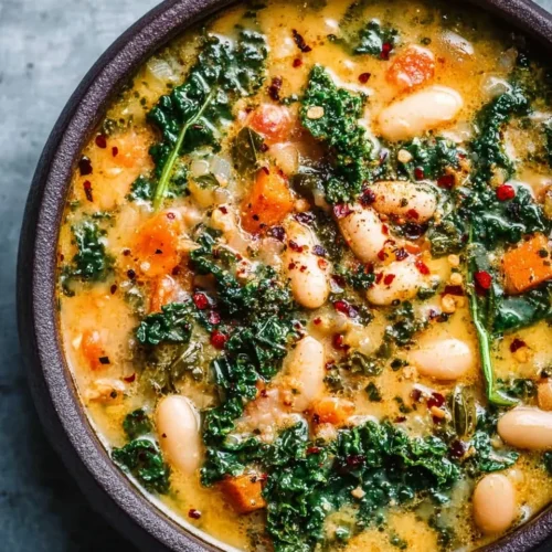 Top-down close-up view of a rustic dark bowl filled with creamy Tuscan white bean and kale soup. The soup features tender cannellini beans, vibrant green kale leaves, and diced carrots in a rich, golden broth, garnished with crushed red pepper flakes.