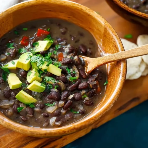 Close-up of thick black bean soup in a wooden bowl with a wooden spoon, topped with fresh diced avocado and cilantro.