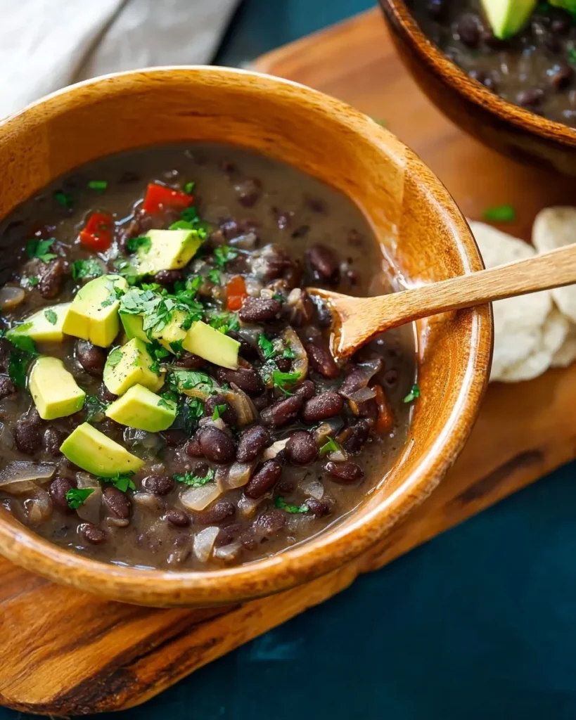 Close-up of thick black bean soup in a wooden bowl with a wooden spoon, topped with fresh diced avocado and cilantro.