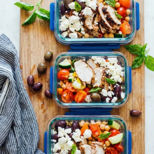 Overhead shot of three glass meal prep containers filled with Mediterranean chicken, chickpeas, tomatoes, cucumbers, olives, and feta cheese arranged on a wooden cutting board with a striped napkin.
