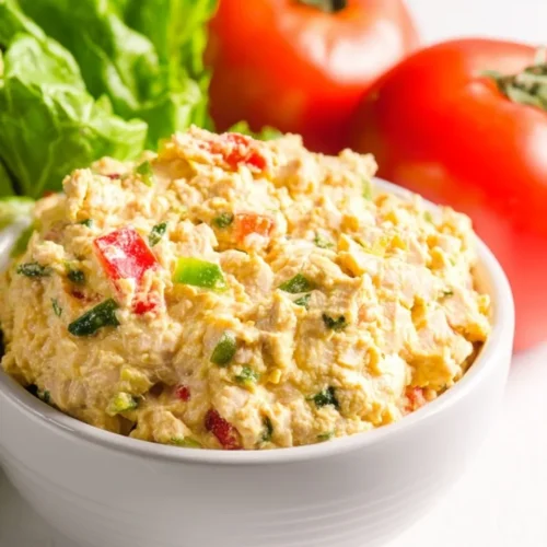 creamy vegan chickpea tuna salad in a white bowl, speckled with diced red peppers and green herbs, with fresh tomatoes and lettuce in the background.