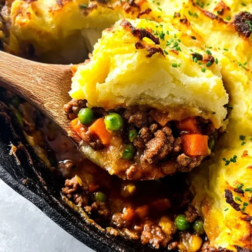 A close-up macro shot of a wooden spoon scooping a generous serving of Beef Stew Shepherd's Pie from a cast iron skillet, revealing layers of savory ground beef filling with carrots and peas underneath a golden-brown, fluffy mashed potato topping garnished with chives.