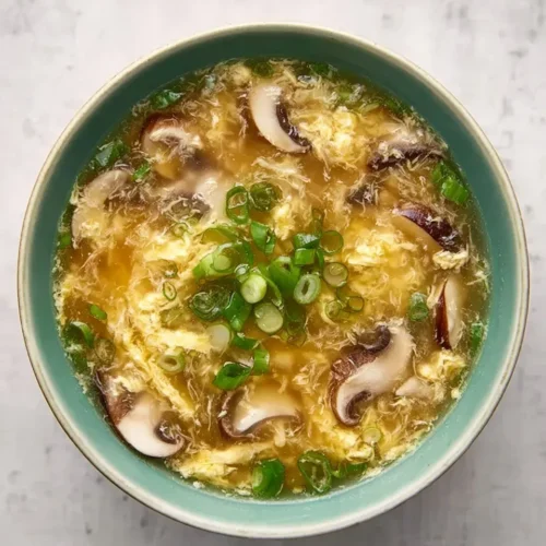Overhead view of a teal bowl filled with hot emergency egg drop soup, featuring silky egg ribbons, sliced shiitake mushrooms, and a fresh garnish of chopped green onions on a light grey background.