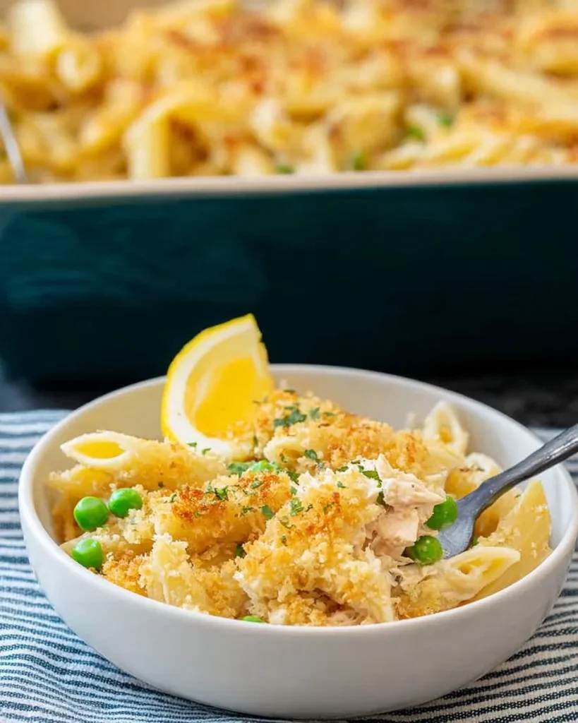 Close-up of a white bowl filled with creamy tuna pasta bake featuring penne, green peas, and a golden breadcrumb topping, garnished with a fresh lemon wedge and a fork ready to take a bite.