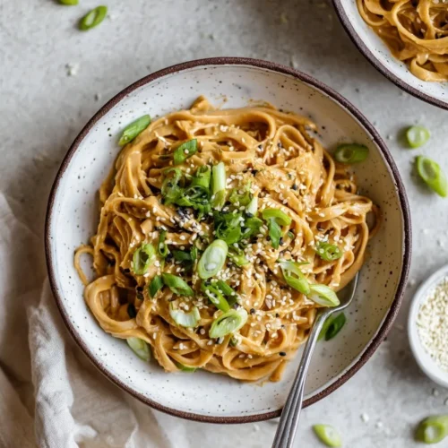 Overhead shot of creamy peanut noodles served in a speckled ceramic bowl, garnished with fresh chopped green onions and sesame seeds, with a silver fork resting in the pasta.
