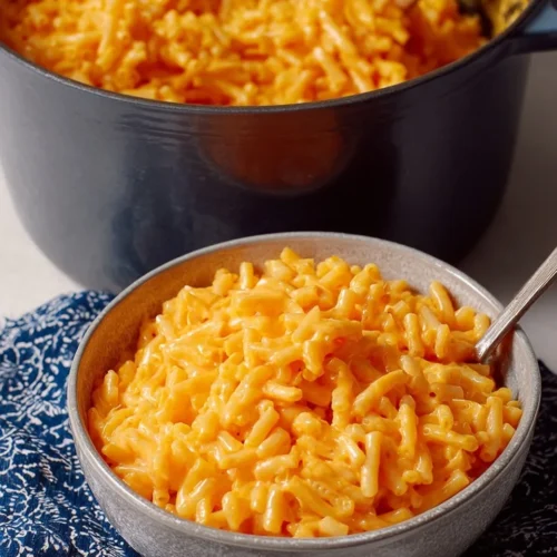 Close-up vertical shot of a grey ceramic bowl filled with creamy upgraded boxed mac and cheese, resting on a blue patterned napkin with a large pot of pasta in the background.
