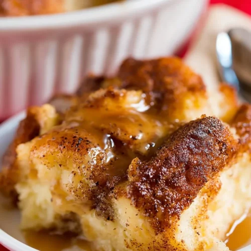 Close-up of a fluffy slice of bread pudding from stale bread on a white plate, generously drizzled with a glossy brown sugar butter sauce, showing the golden cinnamon crust and soft custardy interior.