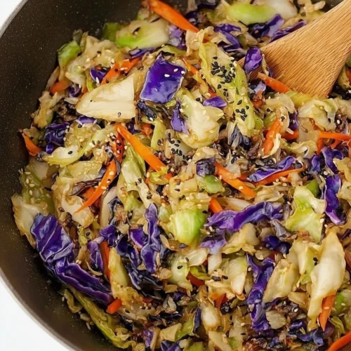Top-down view of a colorful cabbage stir fry cooking in a black wok, featuring shredded green and purple cabbage, carrots, and sesame seeds being tossed with a wooden spatula.