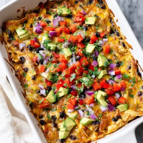 Top-down view of a baked chicken enchilada casserole in a white rectangular baking dish, generously topped with fresh diced avocado, tomatoes, red onion, and chopped cilantro on a marble surface.