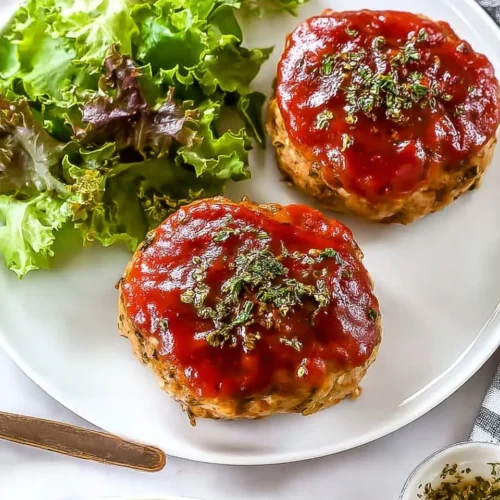 Top-down view of a white dinner plate featuring two glazed mini turkey meatloaves sprinkled with fresh parsley, served next to a fresh green salad.